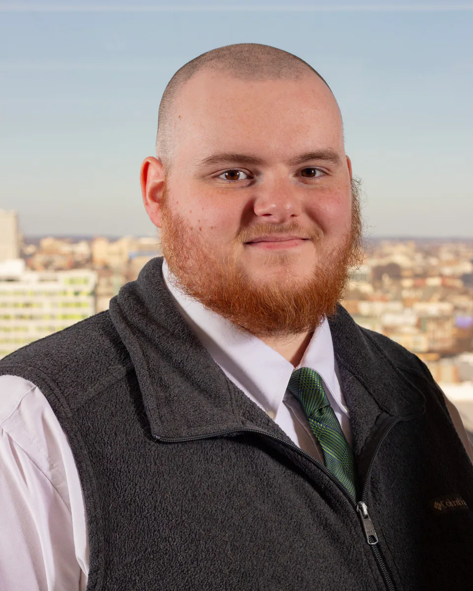 Cole Lyons in a checked shirt and dark vest above a city skyline
